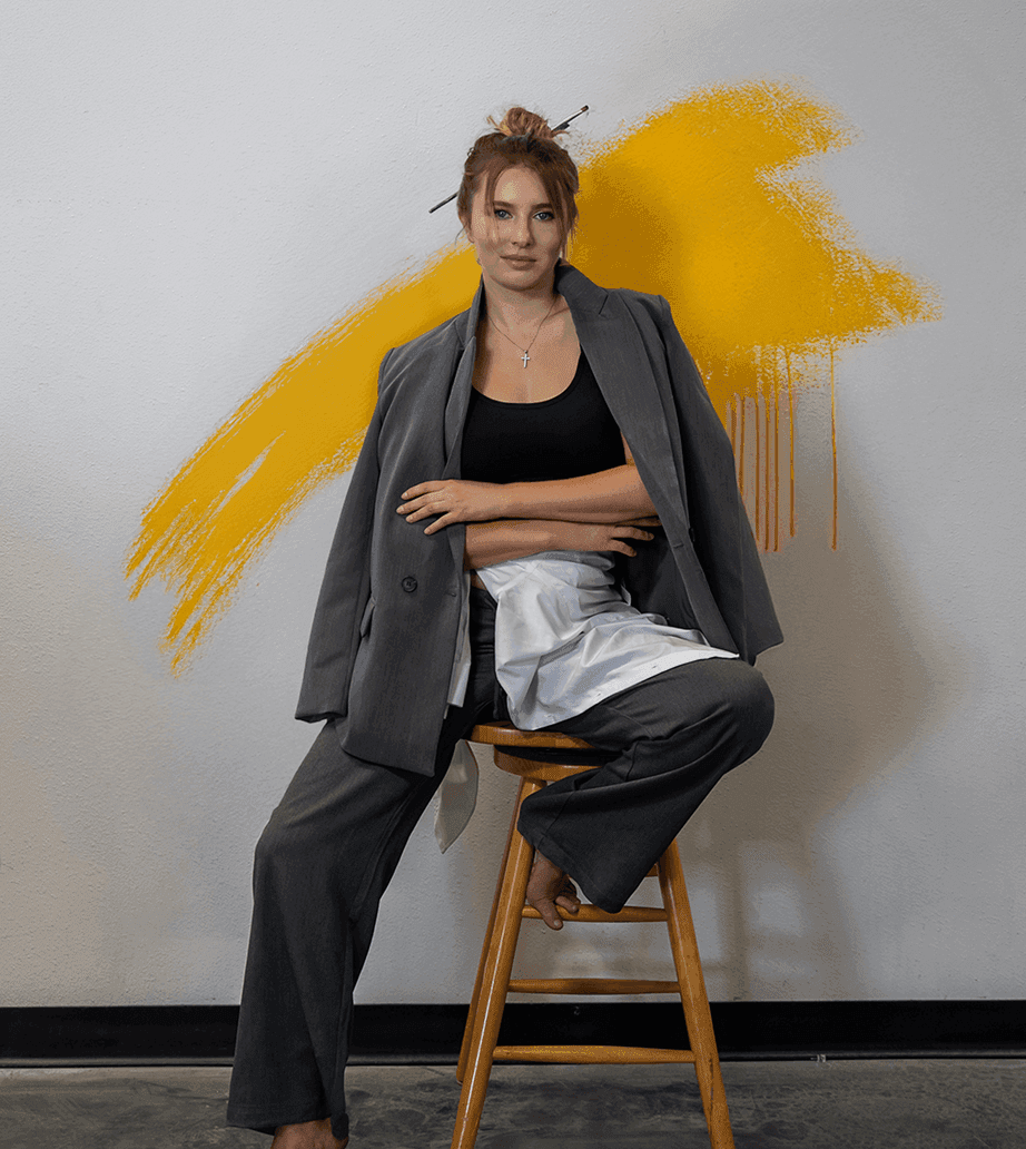 A woman in a blazer and artist's apron sits on a stool with a yellow brushstroke behind her.